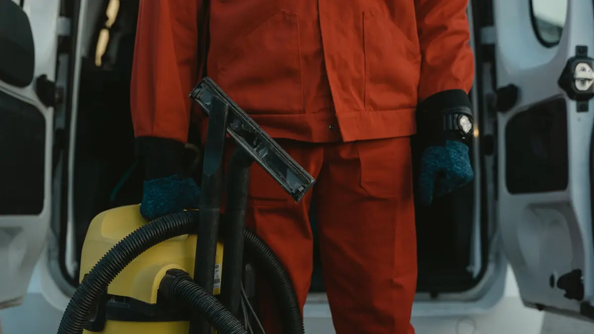 Trabajador de limpieza con uniforme rojo y aspiradora frente a la puerta abierta de una camioneta de servicio